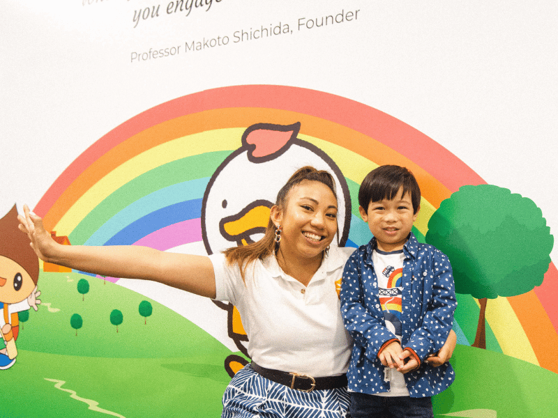 Shichida Australia Promotions - A friendly Shichida teacher smiles alongside a young child. They are in front of a colourful mural featuring a rainbow.