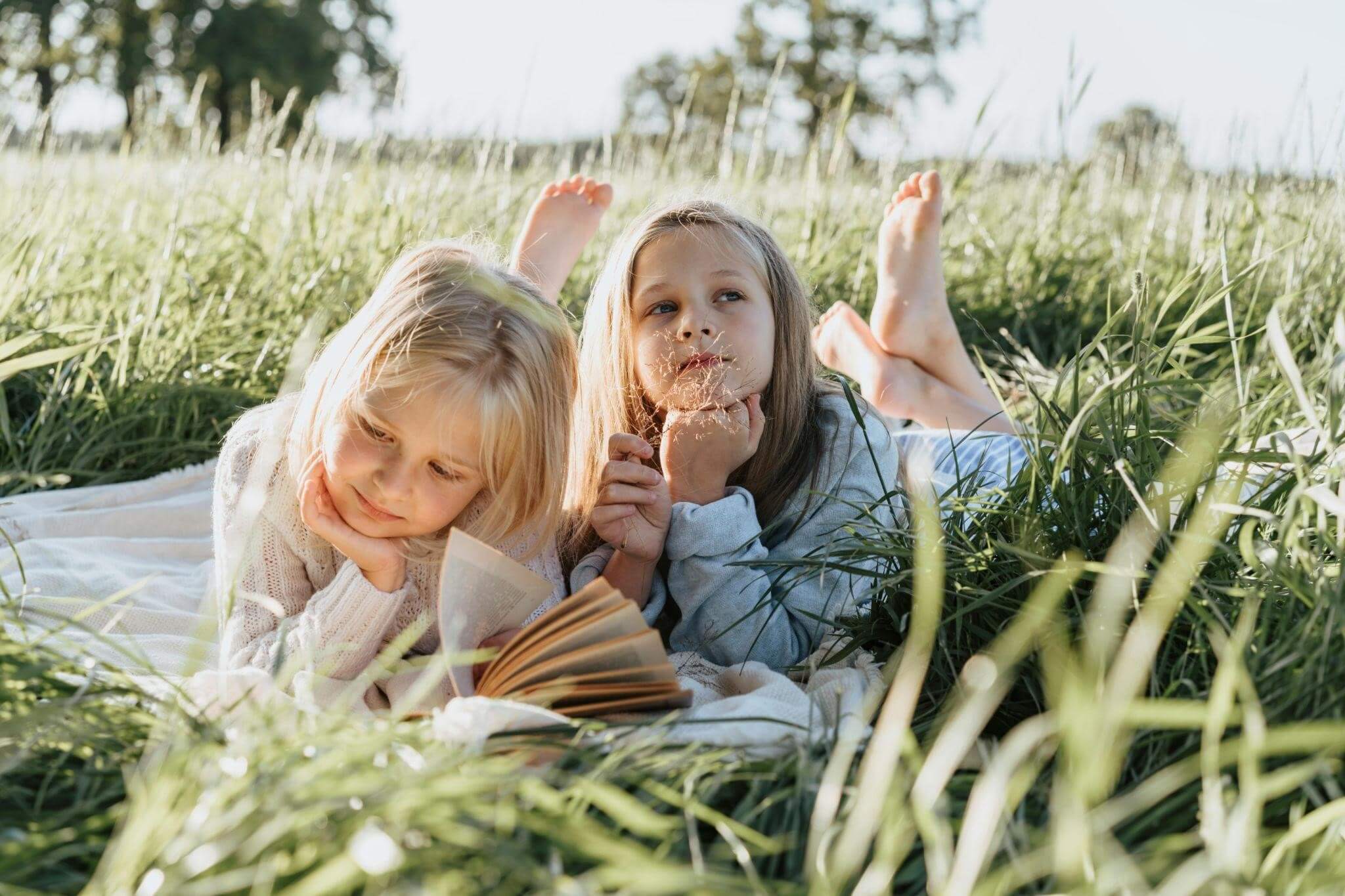 Two girls lying on a blanket in a field of grass, one is reading the other is thinking. This represents critical thinking games for kids