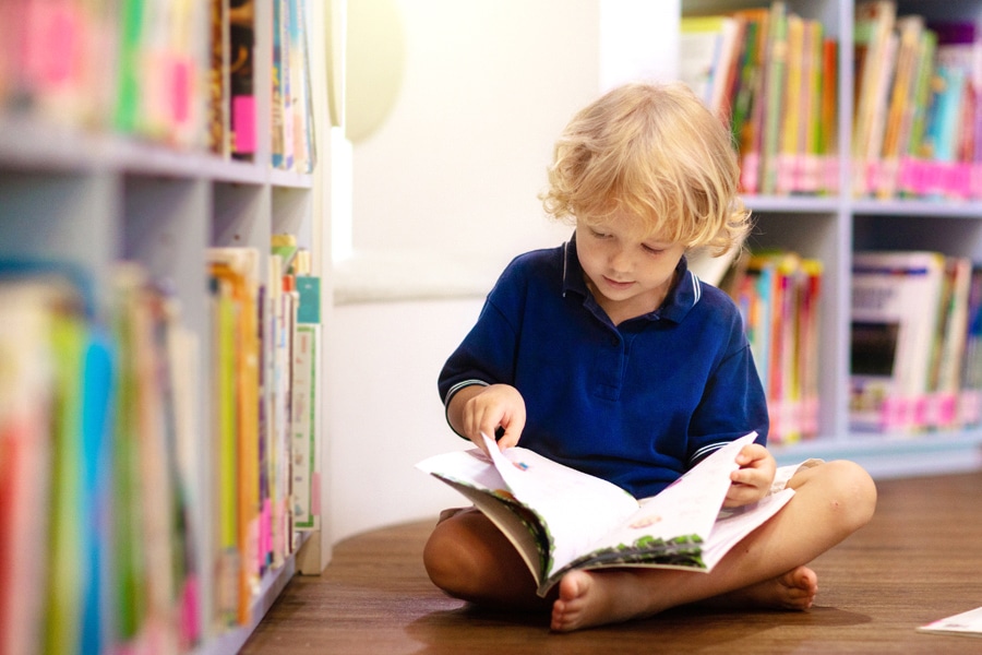 7 benefits of reading for kids, featuring a preschooler sitting on the floor, reading in a library
