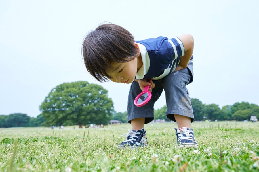 A preschooler shows an inquisitive mind, bending over a field of grass, holding a magnifying glass.