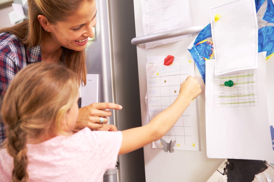 A mum has a visual checklist set up on the fridge, while her preschooler adds stars to tasks she had completed. They are practicing goal setting.