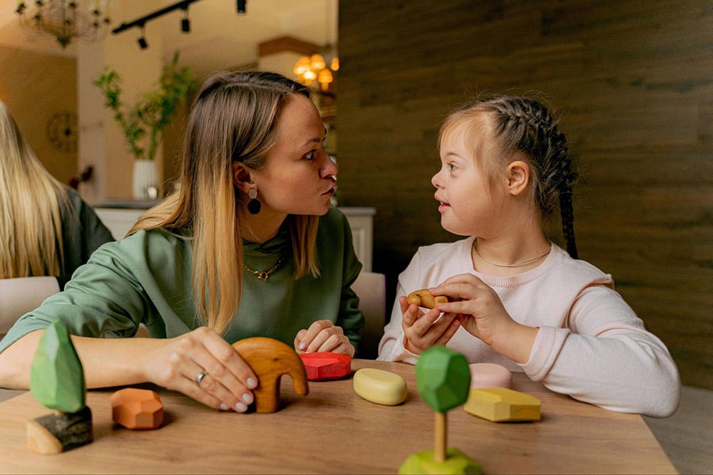 Stages of Child Development Guide, featuring an adult playing with a child who needs extra support. They are sitting at a table playing with wooden toys.