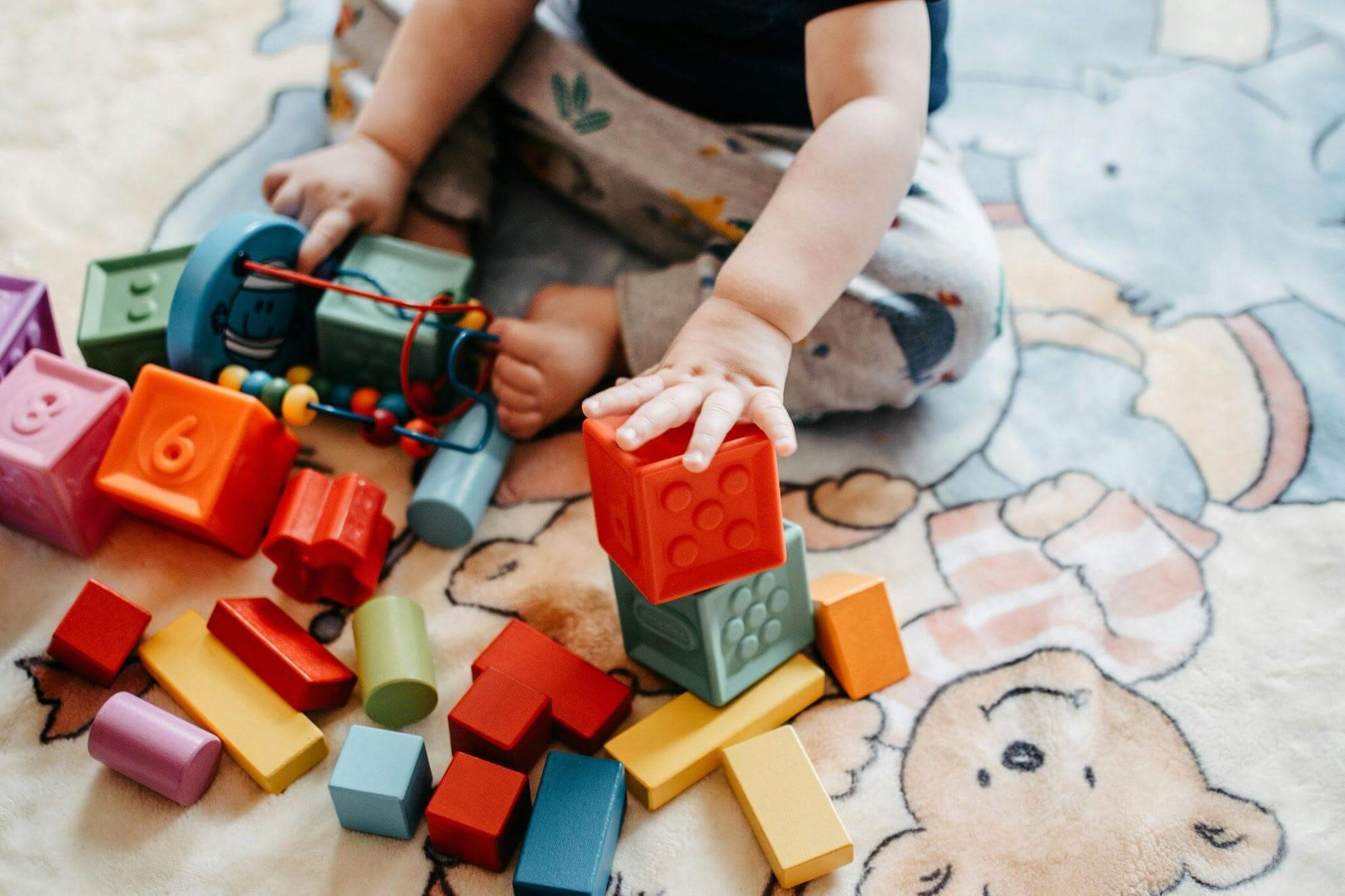 A baby sits on a colorful rug, happily playing with various building blocks scattered around